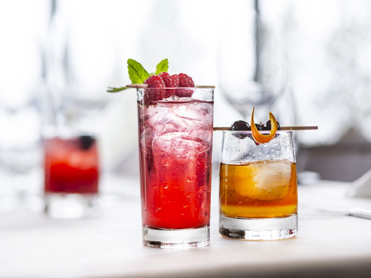 A trio of colorful cocktails on a white table: a tall red drink with raspberries and mint, another red-hued beverage with ice, and a darker amber cocktail with a garnish. Top it at 140 characters, always ending the sentence.