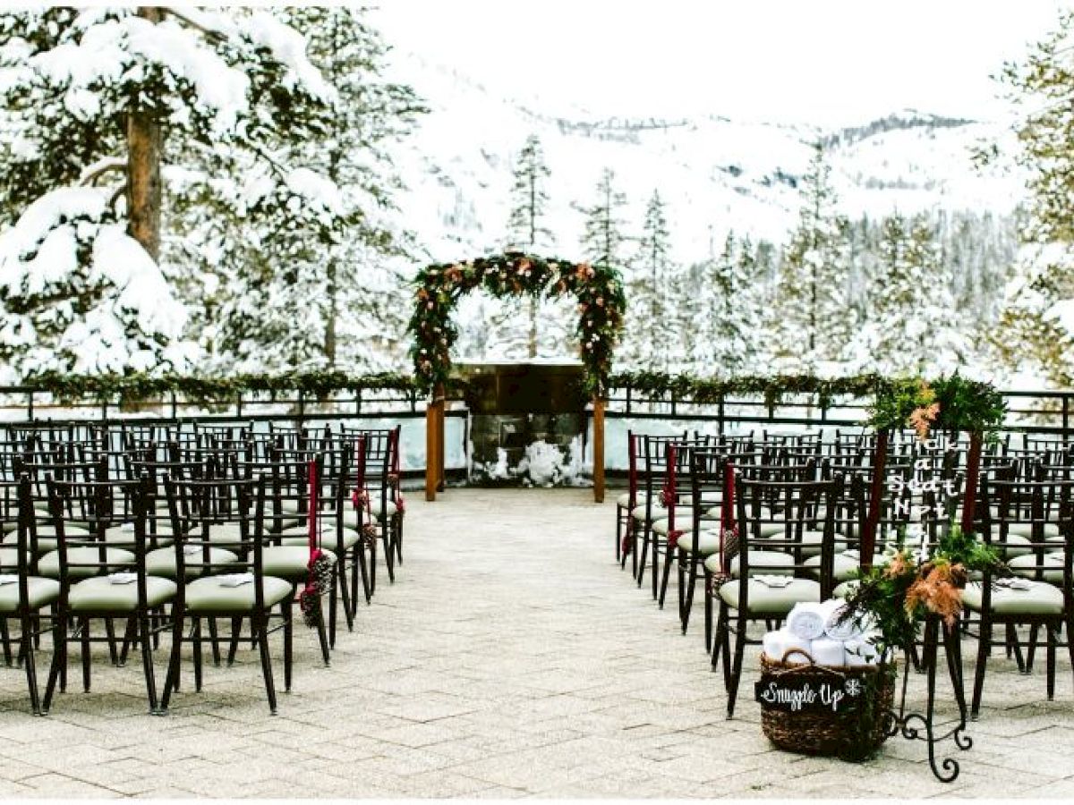 Snowy outdoor wedding setting with chairs, floral arch, and mountainous background, surrounded by snow-covered trees and serene winter scenery.