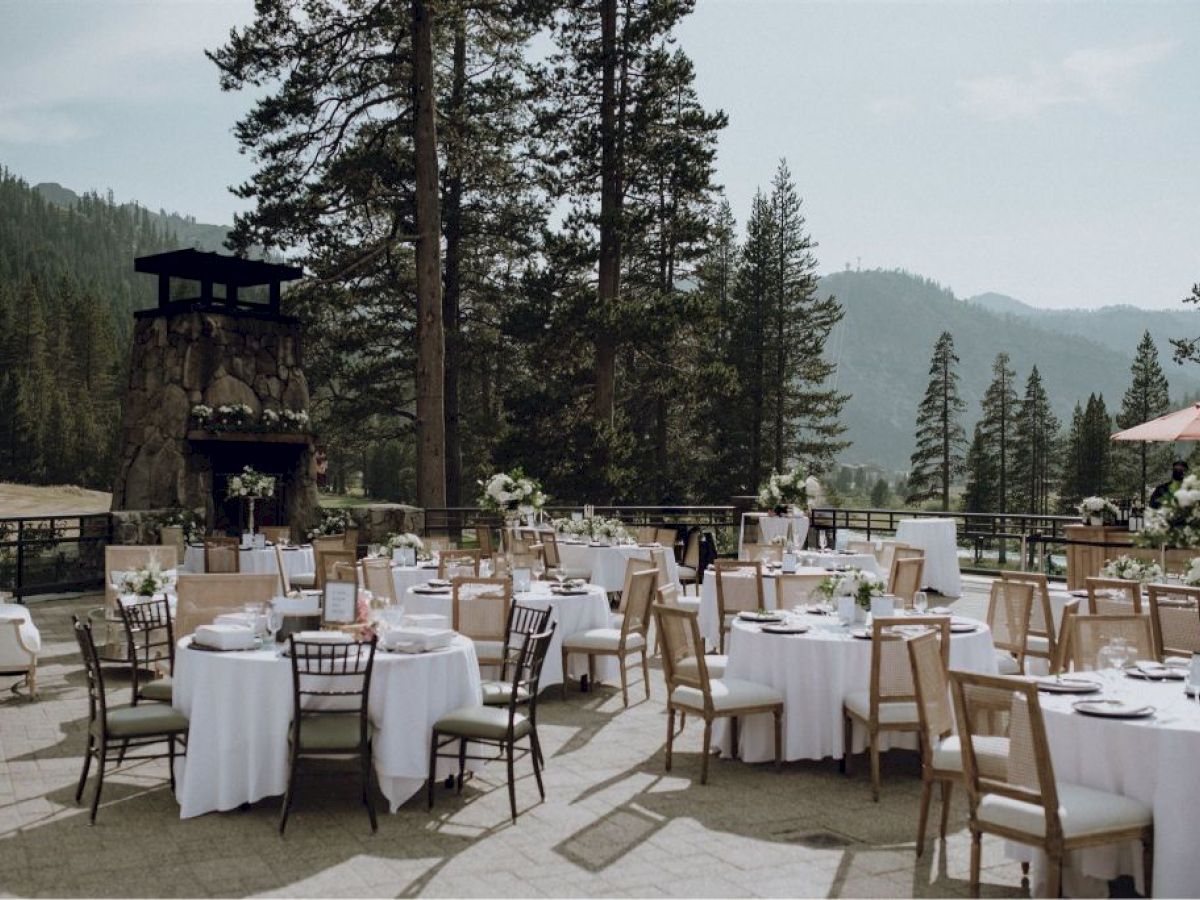 An outdoor event setup with round tables adorned with white tablecloths and floral arrangements, surrounded by a scenic forest and mountains background.