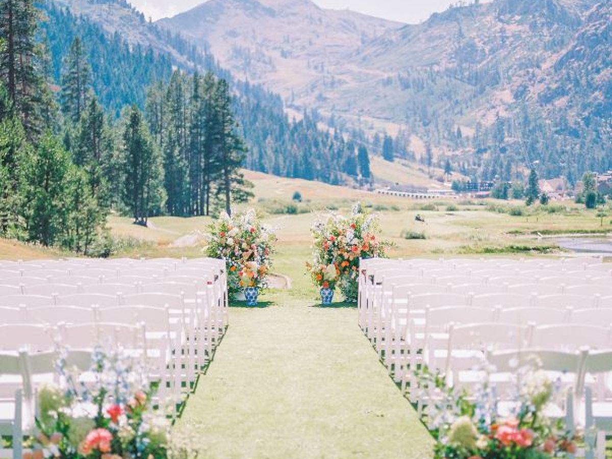 An outdoor wedding setup with rows of white chairs on either side of a grassy aisle, decorated with flowers, mountains, and trees in the background.