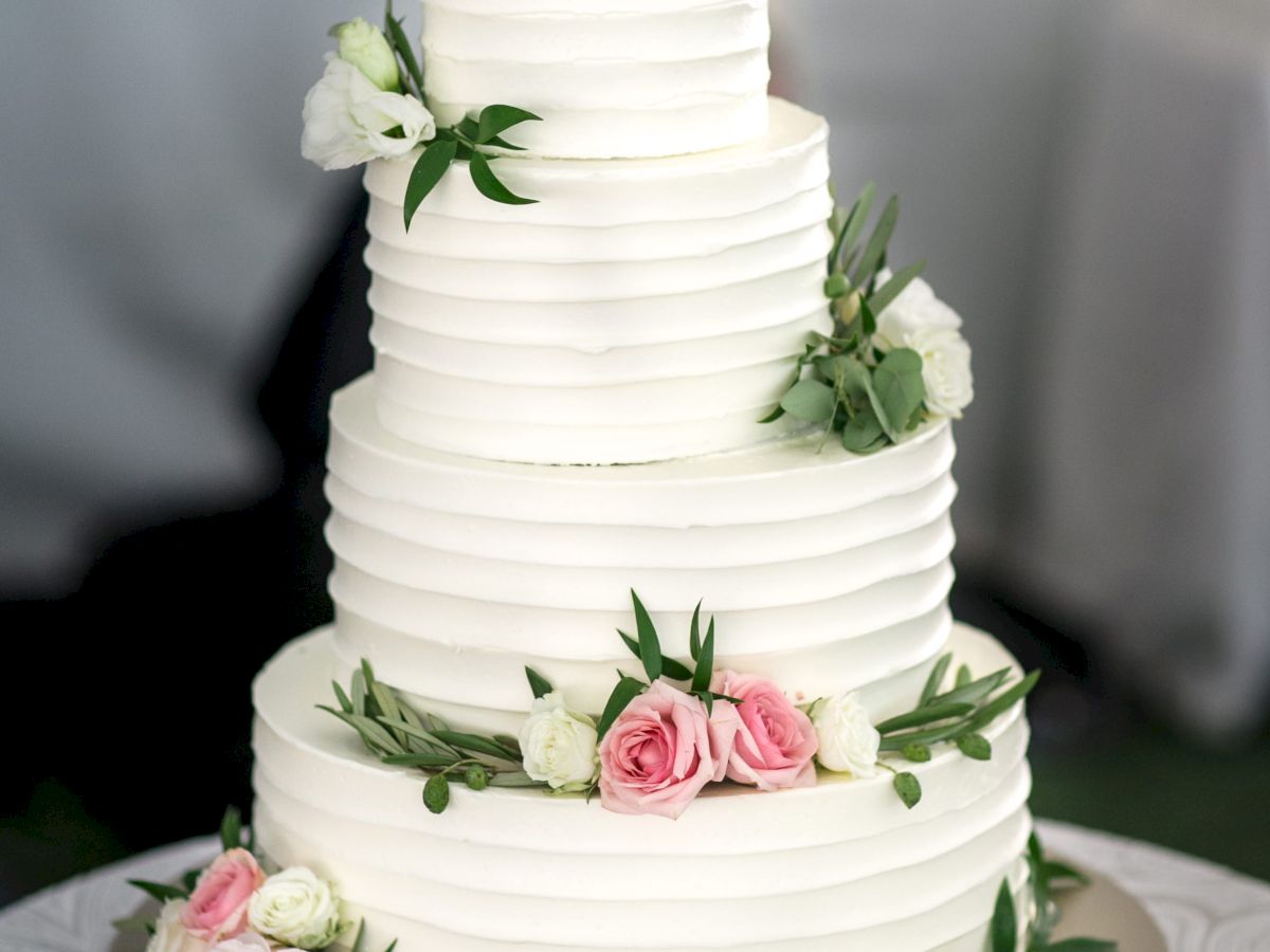 A three-tiered white wedding cake adorned with pink and white flowers and green leaves, placed on a table with plates and a gold cake server.