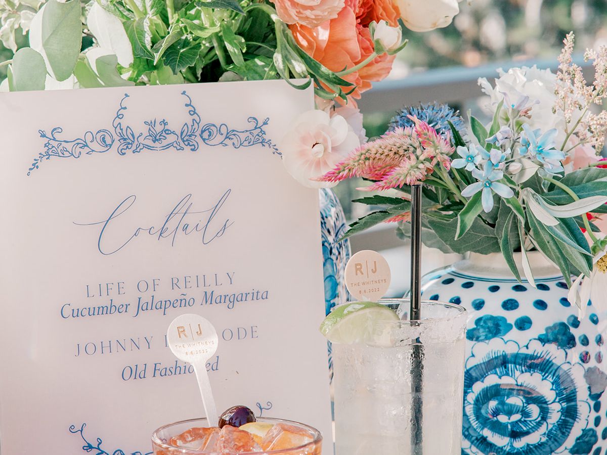 The image shows two cocktails, a floral arrangement, a decorated vase, and a menu card with cocktail names on a table outdoors, potentially at an event.