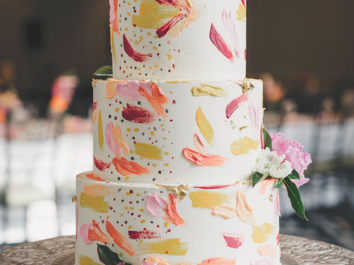 A three-tiered cake with colorful brushstroke decorations and floral accents on top and around, placed on an ornate silver cake stand.