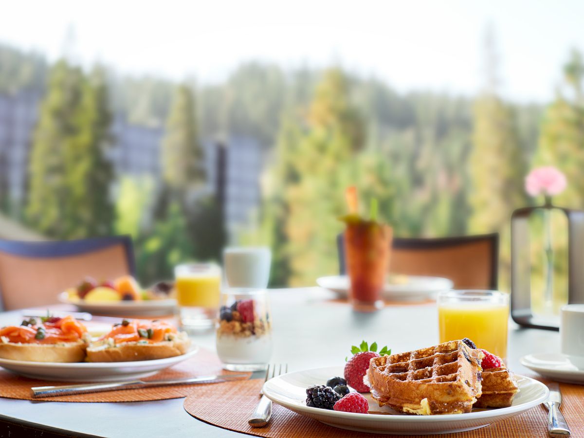 A breakfast table with waffles, juice, fruit, and a parfait, set against a blurry forest view.
