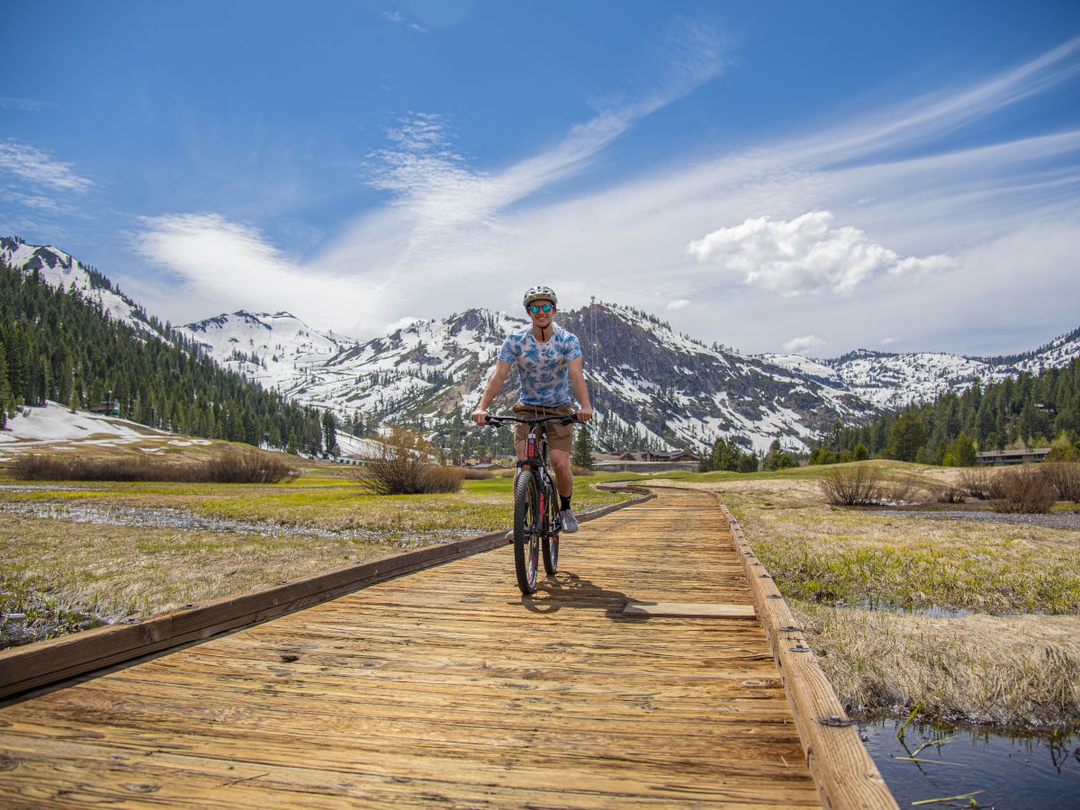 A person is biking on a wooden path through a scenic mountain landscape with patchy snow and a clear blue sky.