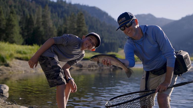 Two people are by a lake, one holding a fishing net and the other holding a fish, both smiling at the camera, surrounded by trees and mountains.