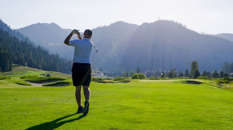 A person is playing golf on a grassy golf course surrounded by mountains and trees under a clear sky with the sun shining brightly.