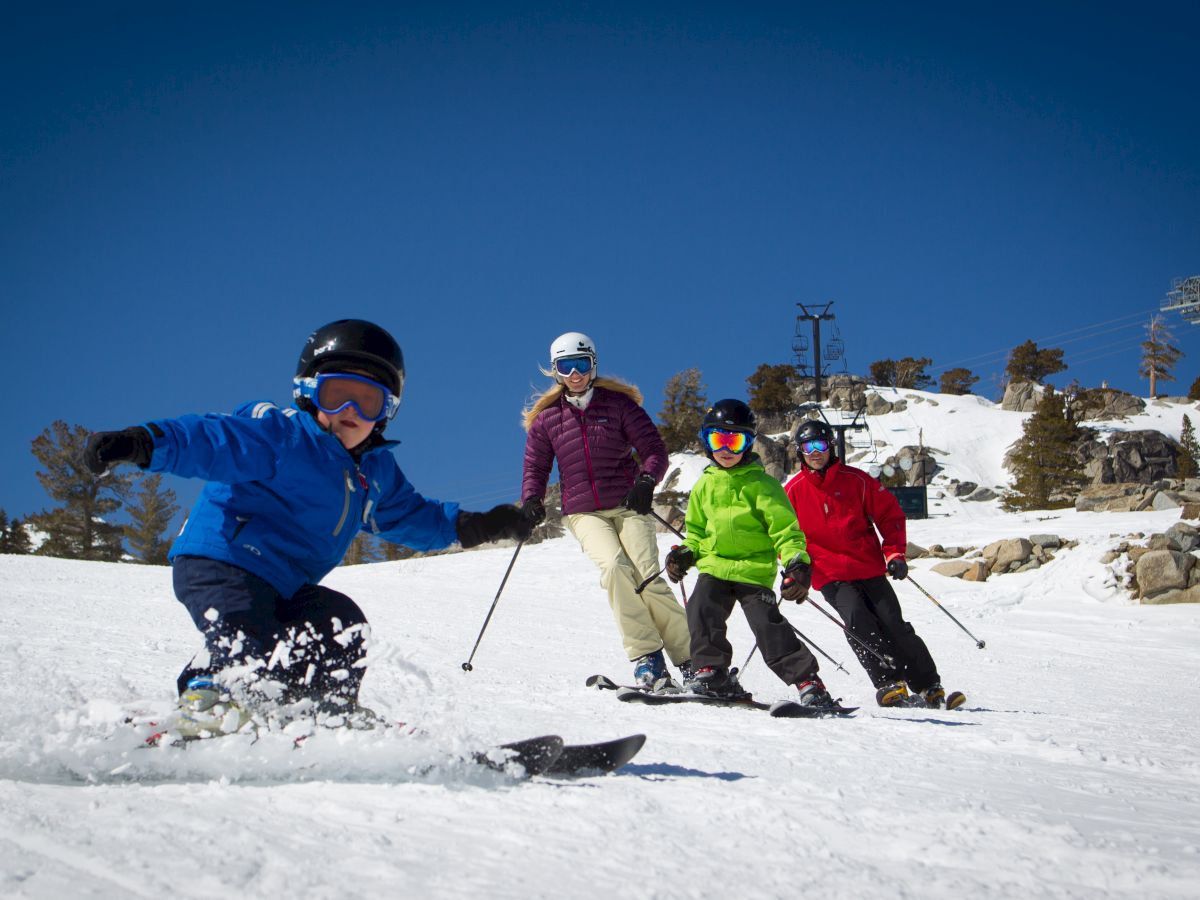 A group of four people skiing down a snowy slope under a clear blue sky, wearing colorful jackets and helmets.