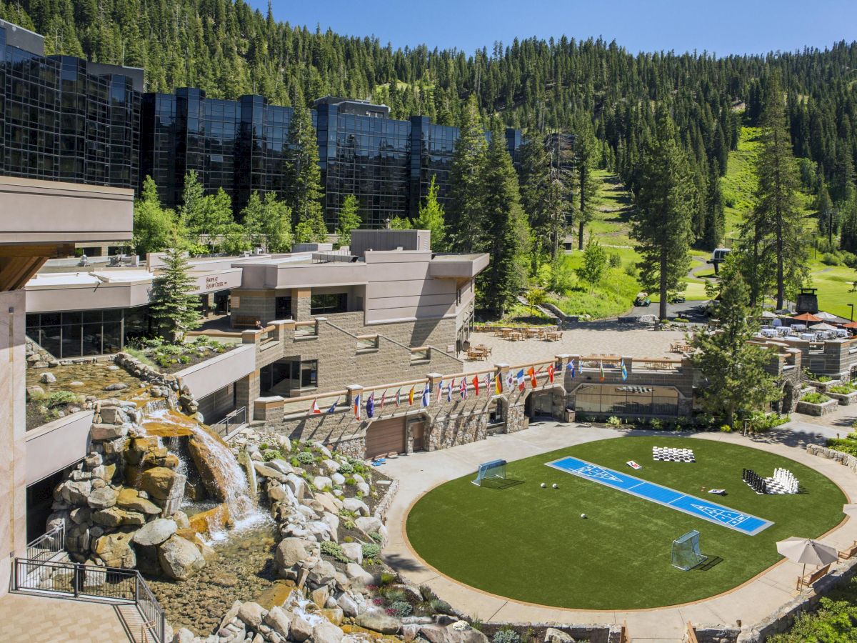 A picturesque resort surrounded by lush green forest, featuring modern buildings, a waterfall, flag decorations, and a central grassy area.