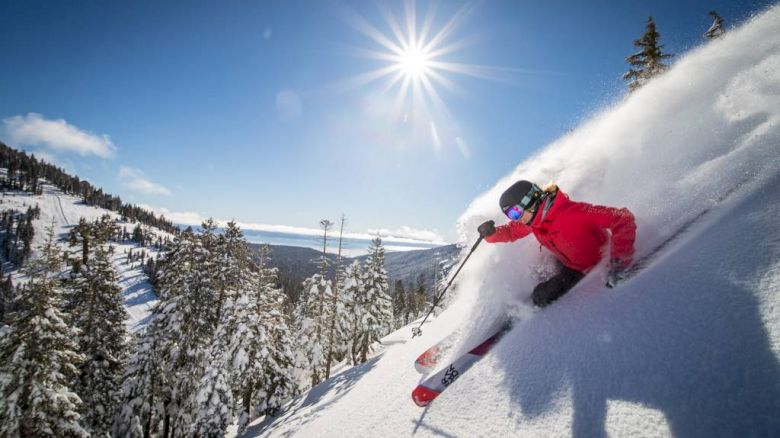 A skier in a red jacket is skiing down a snowy slope, creating a spray of snow under a bright sun, with a scenic mountain background.