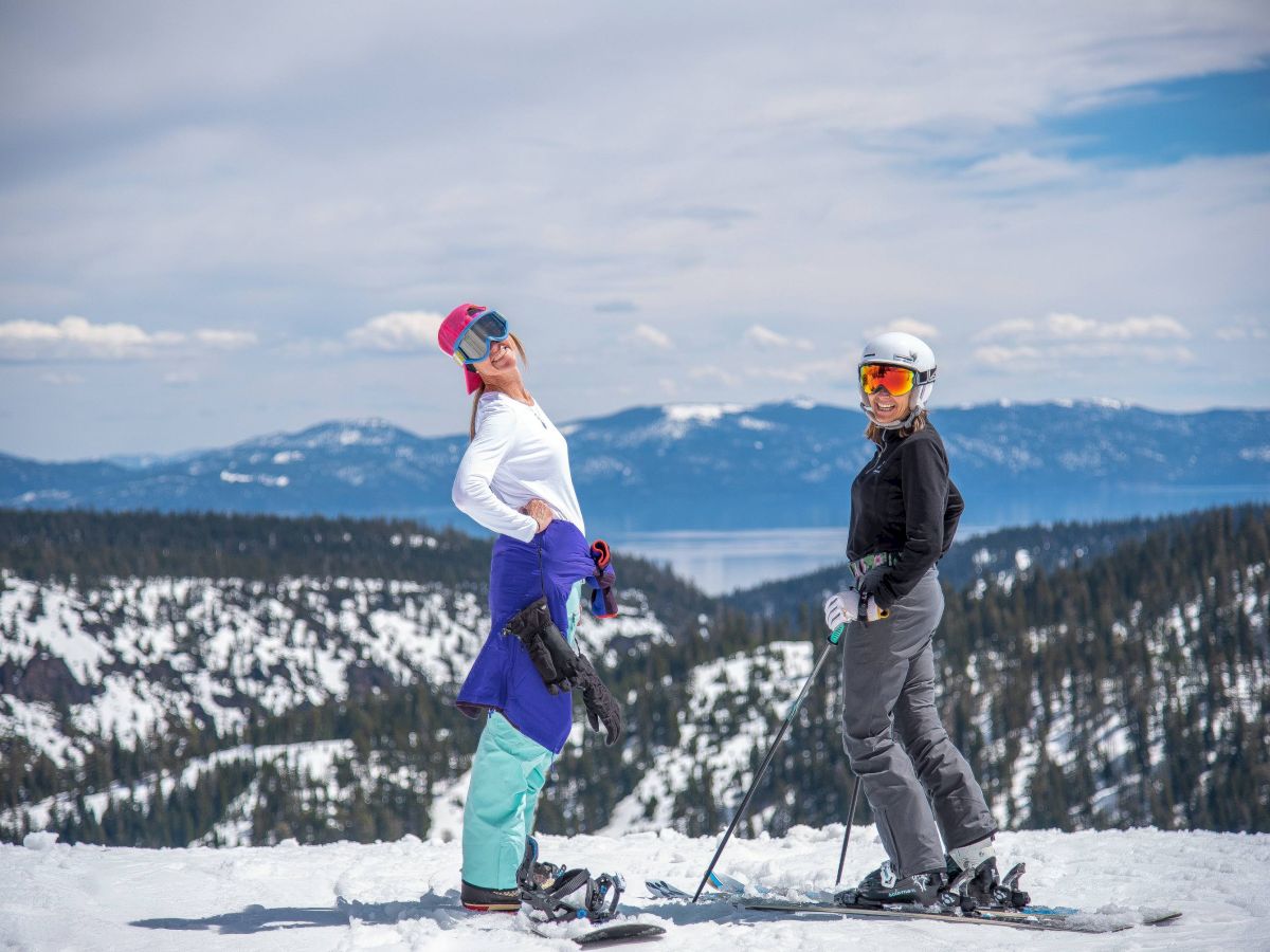 Two people in ski gear posing on a snow-covered mountain with scenic views of trees and mountains in the background.
