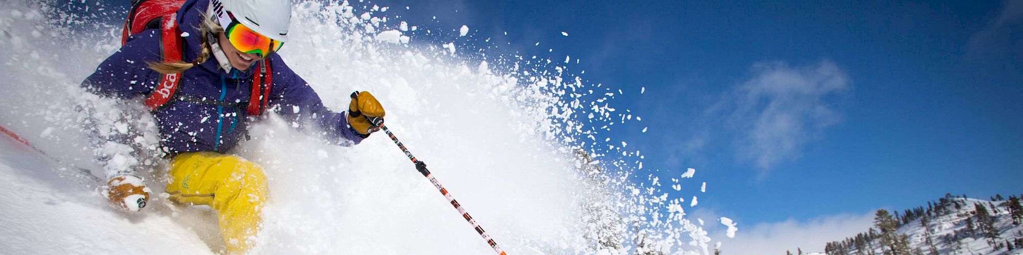 A skier is captured in mid-action, descending a snowy slope with powder flying around, under a clear blue sky.