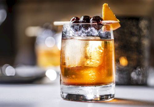 A short, stylish whiskey cocktail on the rocks with an orange twist and berries garnish, served in a chunky glass.