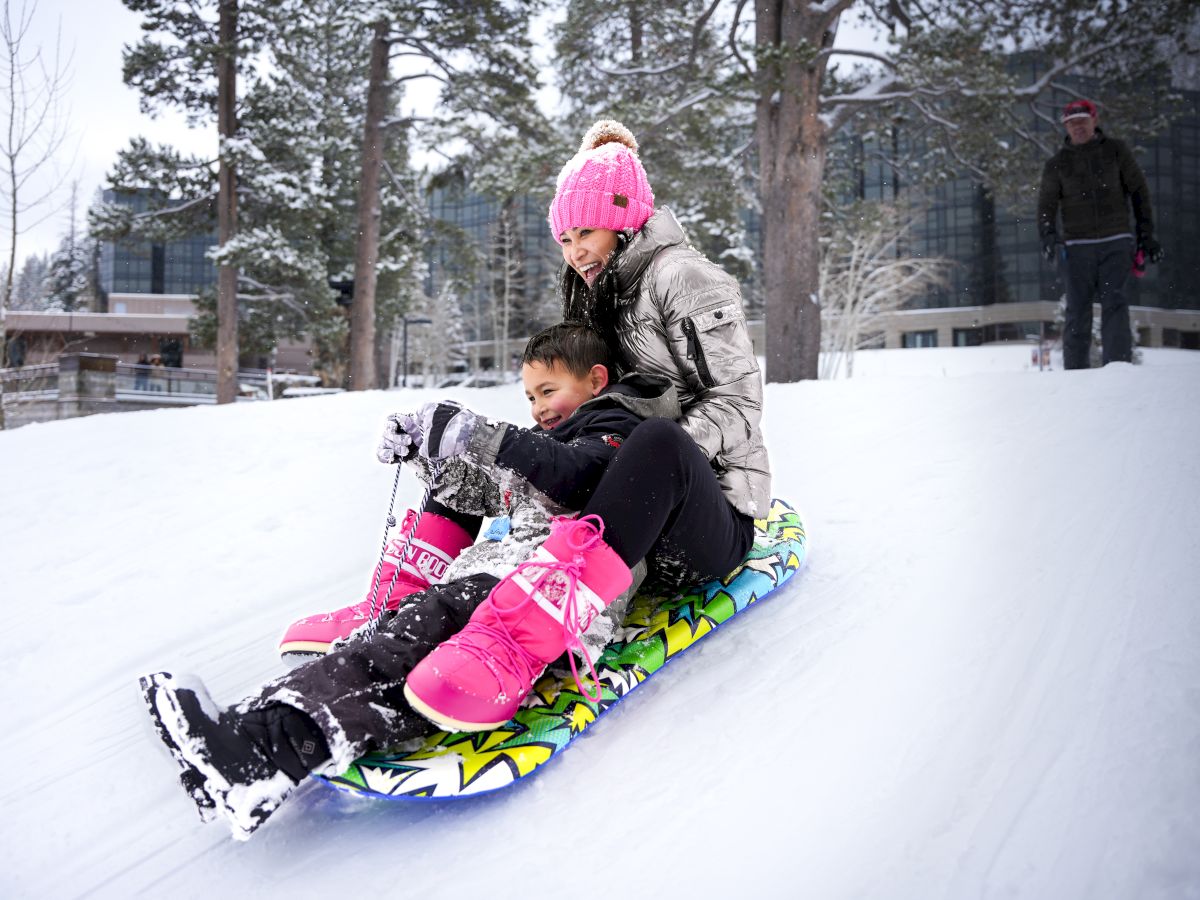 Two kids sledding down a snowy hill with one guiding from behind; a person in a pink hat supervises.