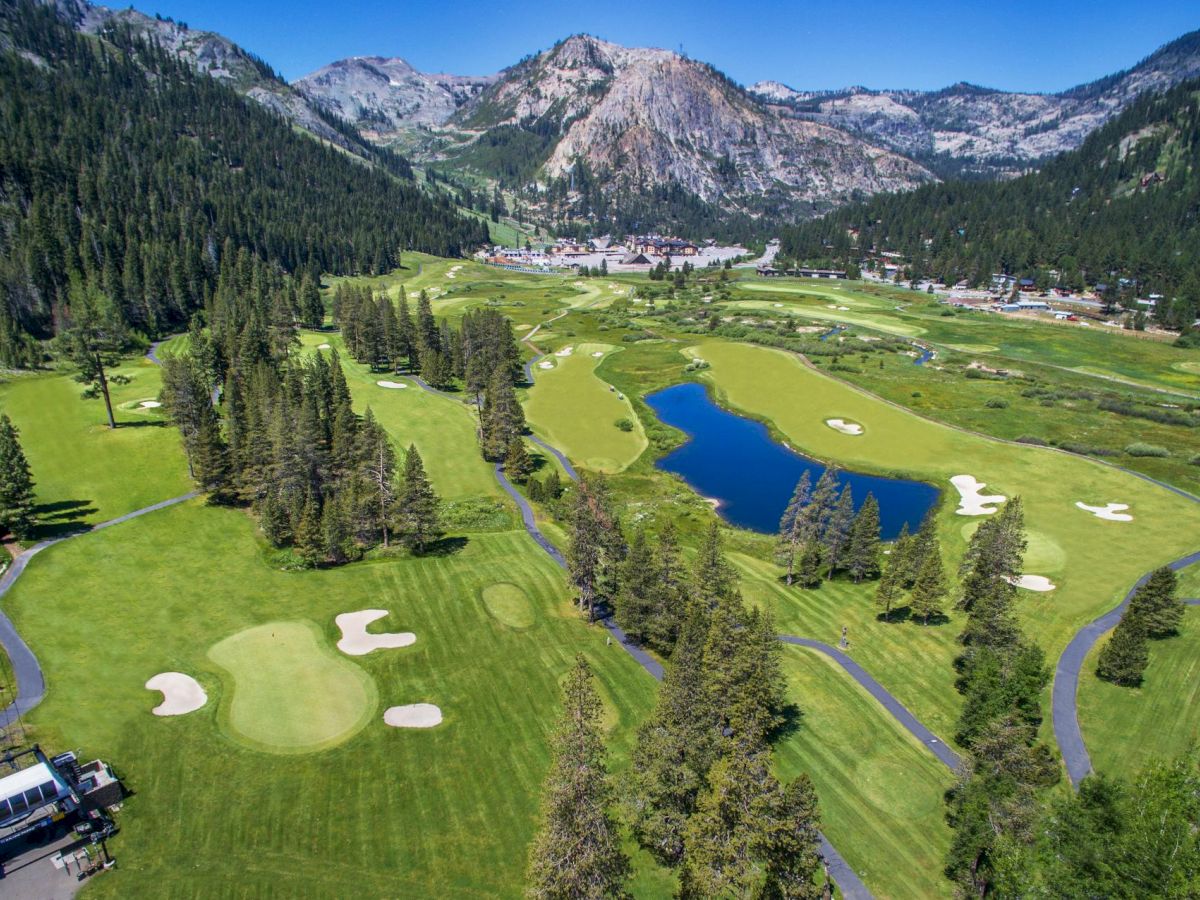 A picturesque golf course situated in a mountainous landscape, featuring greens, sand bunkers, a winding path, a pond, and lush trees.
