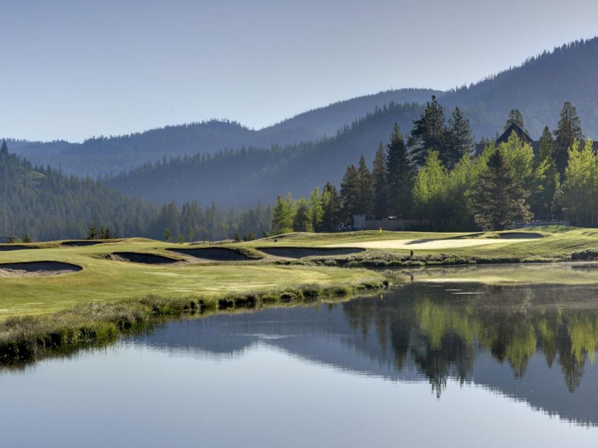 A picturesque golf course with a water hazard, surrounded by forested hills and mountains, and a reflection of trees in the water.