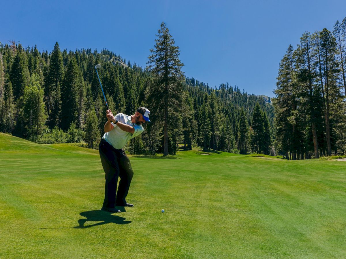 A golfer is mid-swing on a lush, green course surrounded by tall trees and scenic mountains under a clear, blue sky.