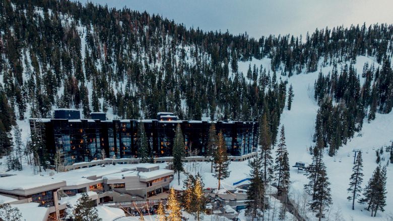 A snowy mountain resort with outdoor pools, surrounded by dense evergreen trees. The buildings are nestled within the snowy landscape and forest.