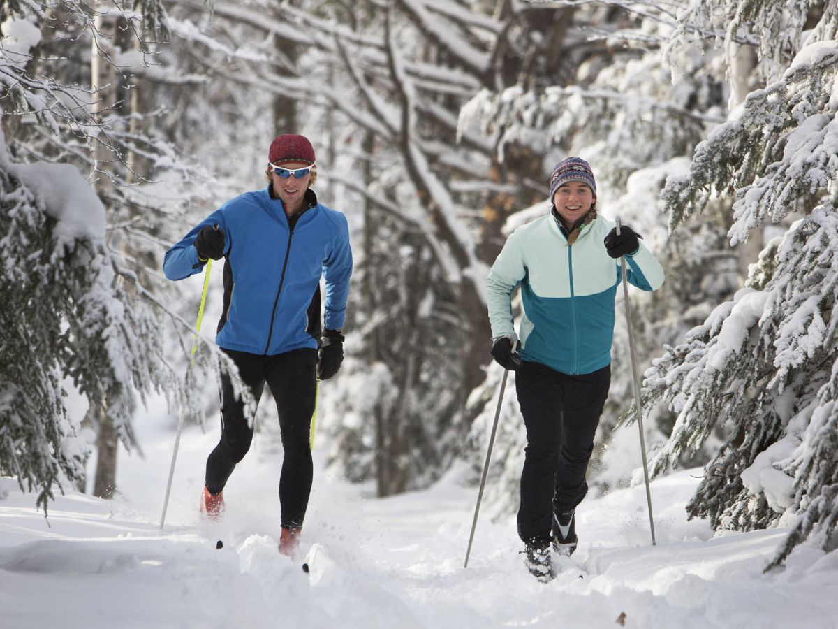 Two people cross-country skiing on a snow-covered trail in a forest, dressed in winter gear and smiling.