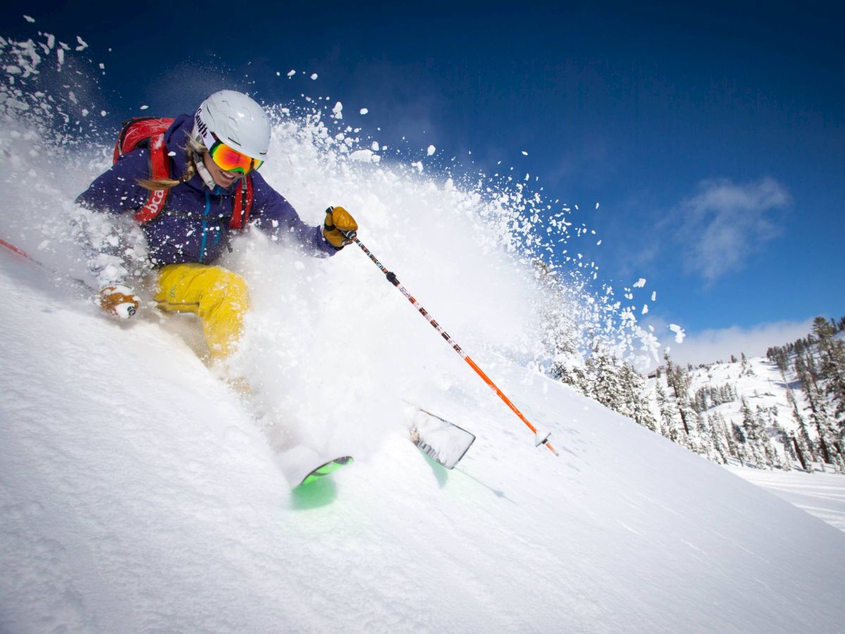A person is skiing down a snowy slope, creating a spray of powder snow, with a clear blue sky and a mountainous backdrop in the distance.