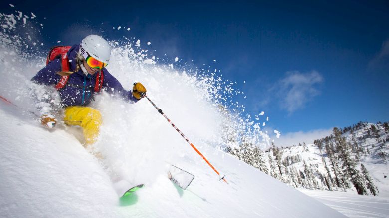 A person is skiing down a snowy slope, creating a spray of powder snow, with a clear blue sky and a mountainous backdrop in the distance.