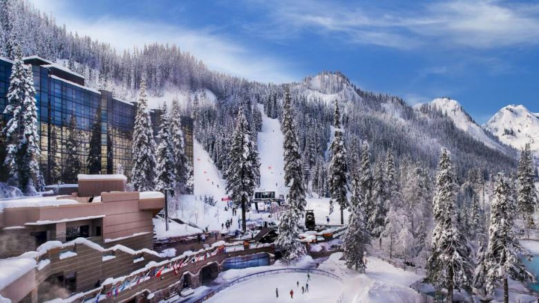 A beautiful winter scene with snowy mountains, a ski slope, a lodge, and people skating on a frozen pond surrounded by trees and buildings.