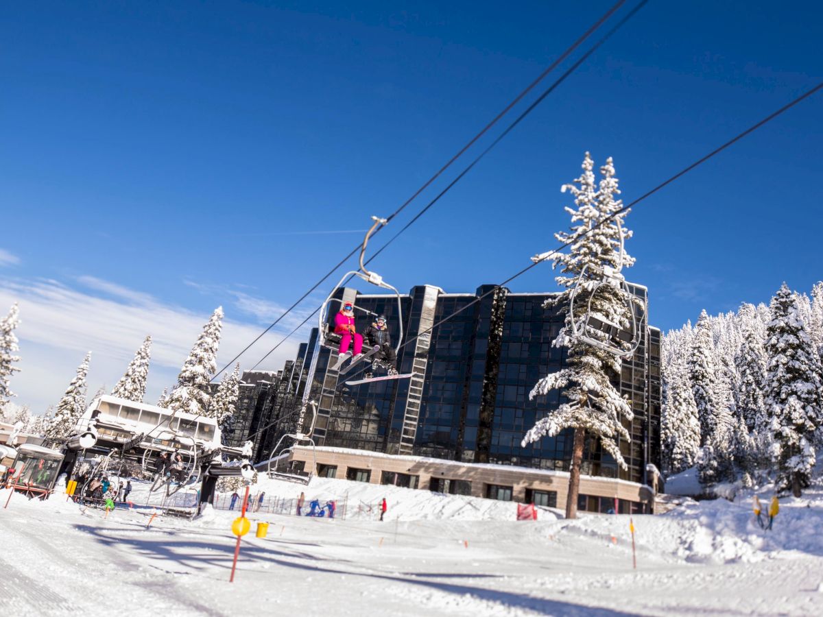 The image shows a snowy ski resort with people on a ski lift, a large building, and snow-covered trees under a clear blue sky.