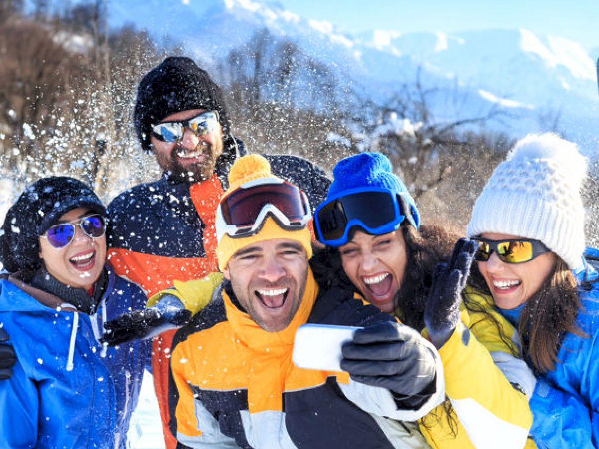 A group of friends in winter gear, having fun in a snowy mountain setting, taking a selfie and smiling cheerfully under the bright sun.