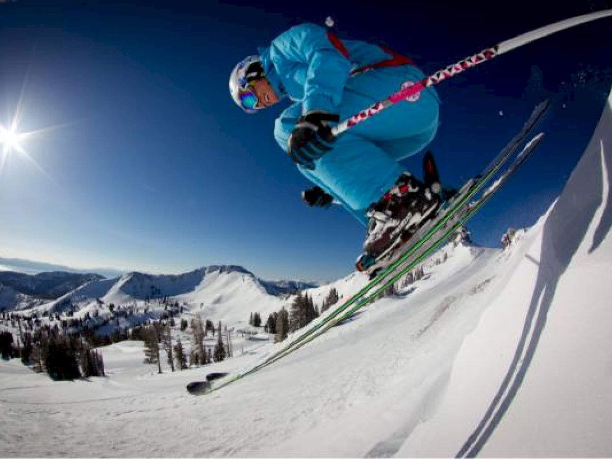 A skier in a blue outfit is airborne, jumping off a snowy slope, with a clear blue sky and mountainous landscape in the background.
