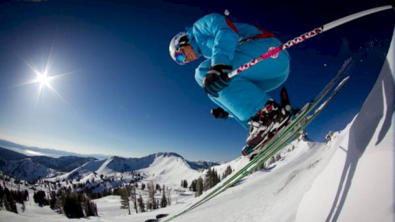 A skier in a blue outfit is airborne, jumping off a snowy slope, with a clear blue sky and mountainous landscape in the background.