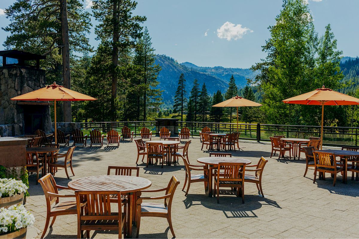 An outdoor seating area with wooden tables, chairs, and orange umbrellas is set against a scenic mountain view with tall trees and a clear sky.