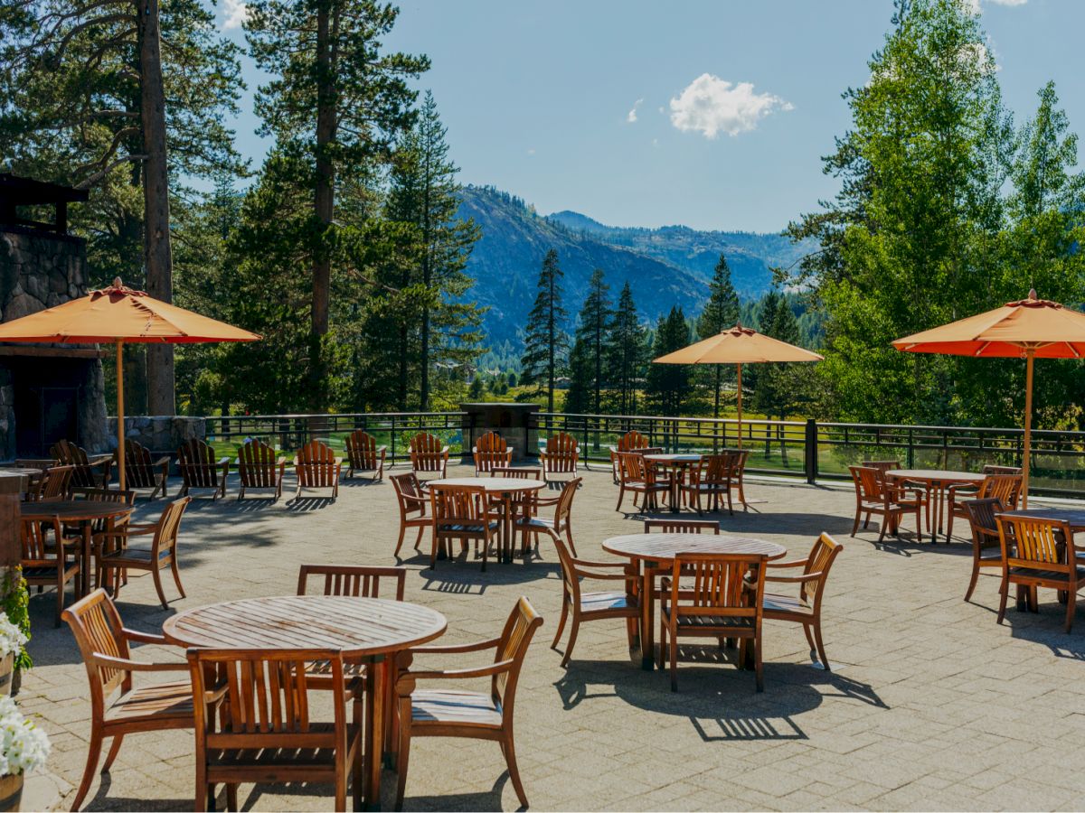 An outdoor seating area with wooden tables, chairs, and orange umbrellas is set against a scenic mountain view with tall trees and a clear sky.