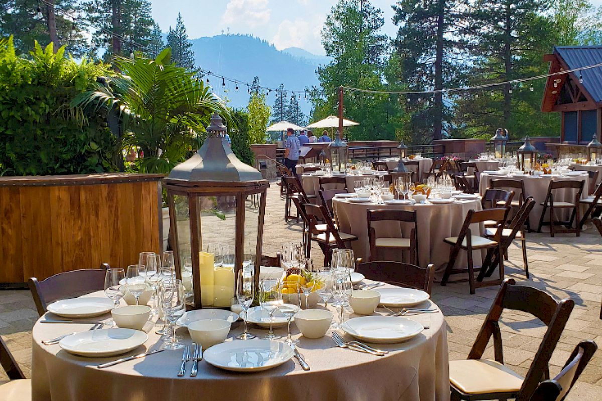 An outdoor event setup with round tables, chairs, and table settings, surrounded by trees and mountains in the background.