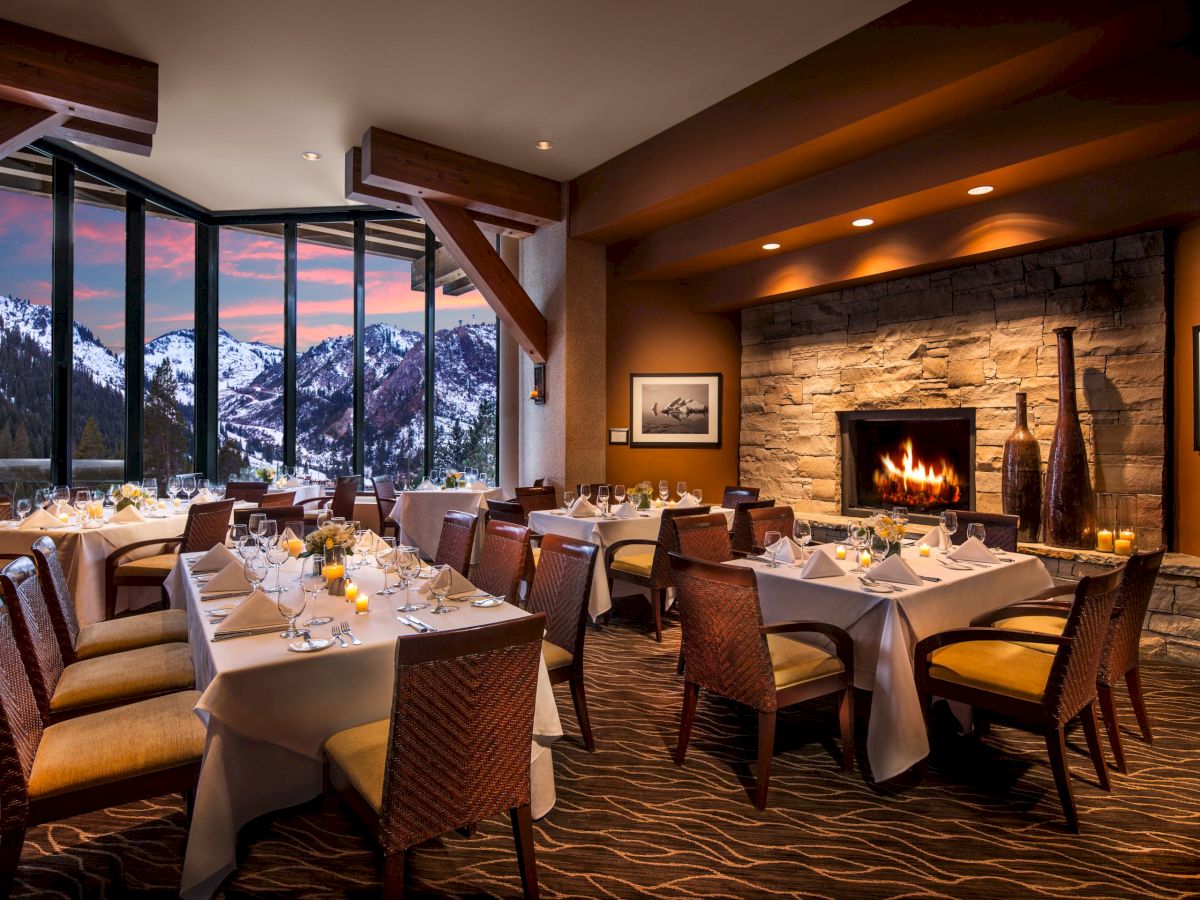 A cozy dining room with a large fireplace, elegantly set tables, and large windows showcasing a snowy mountain view at sunset.