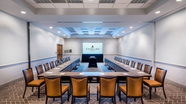 A conference room with a U-shaped table setup, chairs, notepads, and a projection screen displaying 