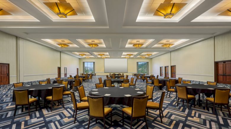 The image shows a large conference room with round tables and yellow chairs, patterned carpet, modern ceiling lights, and a projection screen.