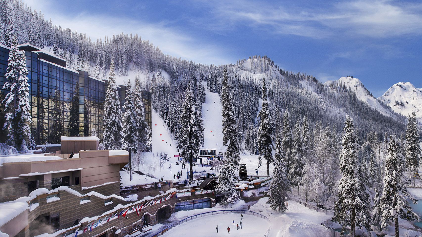 A snowy mountain resort with ski slopes, tall trees, buildings, and a blue sky with scattered clouds in the background, surrounded by snow-covered peaks.