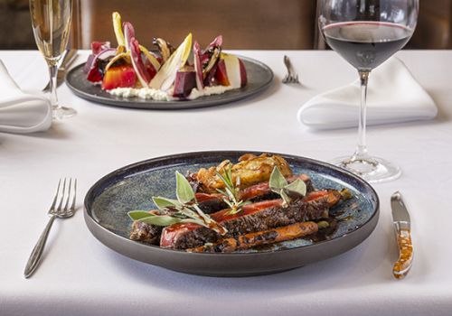 A fancy plated steak with greens and garnishes on a dark ceramic plate, on a white-tablecloth setup with wine glasses in the background.