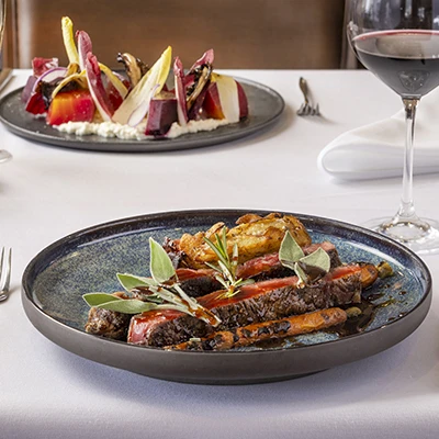A fancy plated steak with greens and garnishes on a dark ceramic plate, on a white-tablecloth setup with wine glasses in the background.
