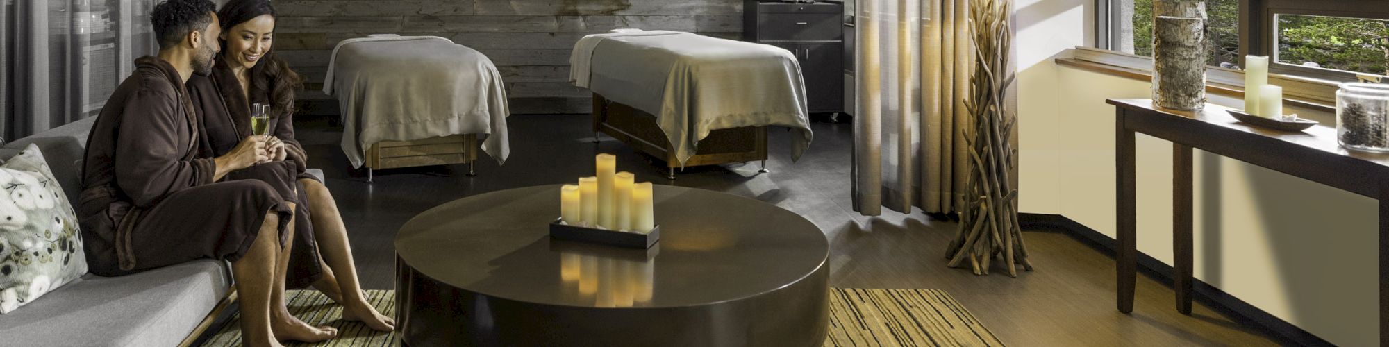 The image shows a serene spa room with a couple in robes seated together, massage tables, candles, large windows, and natural decor elements.