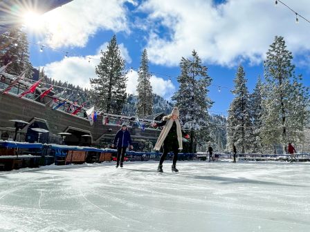 People skating on a snowy outdoor rink with tall pine trees, blue sky, and a sunburst; cozy arena vibe and festive lights at the top.