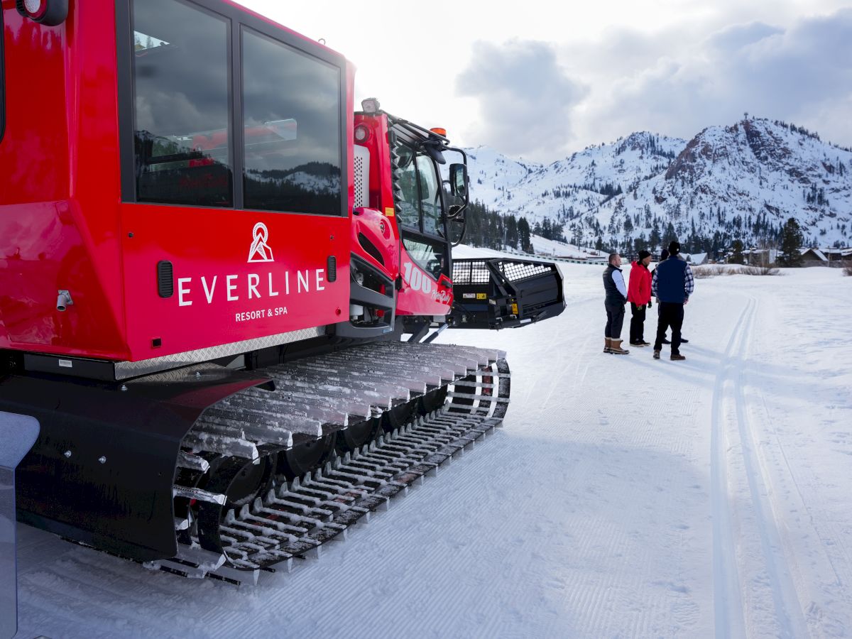 Red snowcat machines labeled EVRLINE on a snowy slope, with mountains and a few people in the background.