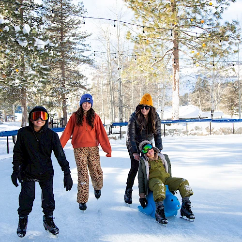 A group of four kids in winter gear skate on a snowy outdoor rink, with trees, string lights, and a fence in the background.