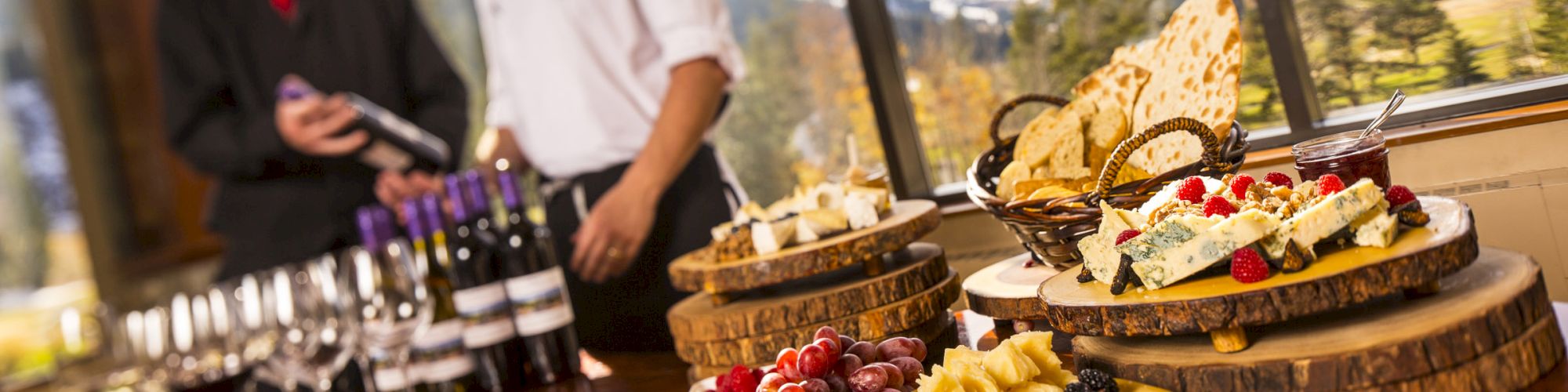 A table with wine bottles, glasses, and plates of cheese and fruit; two people in the background, nature and mountains visible outside the window.