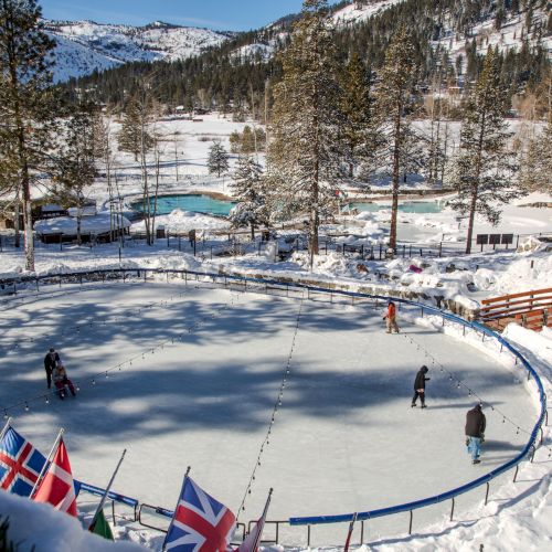 Snowy mountain ski resort with skiers on a fenced slope, Union Jack flags, forested valley, and hills under bright blue sky.