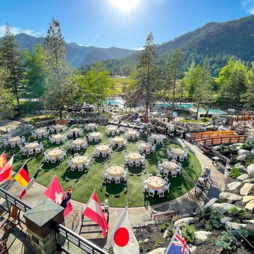 A mountain resort patio set for a banquet: round white chairs, tables, flags, and a pool amid green valleys.