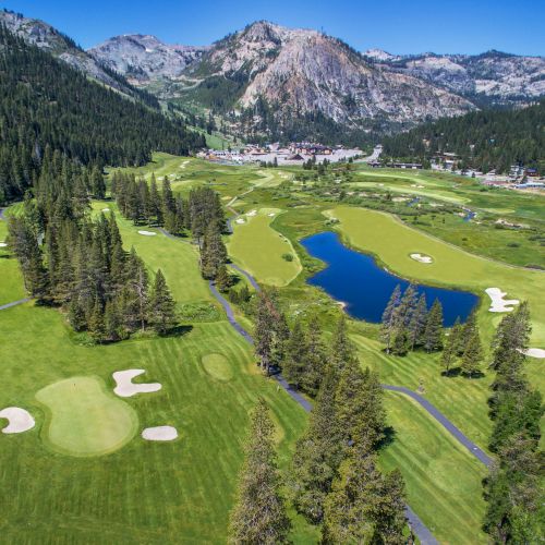 An aerial view of a lush golf course with a winding pond, manicured fairways, cabins, and dramatic alpine mountains in the background.
