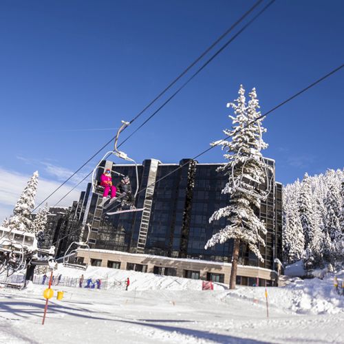A snowy ski slope with pine trees, a chairlift, and a large mountain lodge building against a bright blue sky.
