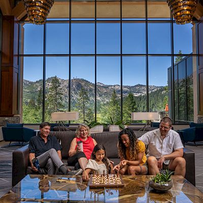 A family sits in a bright lodge living room by a glass table playing a board game, with huge mountain-view windows behind them.