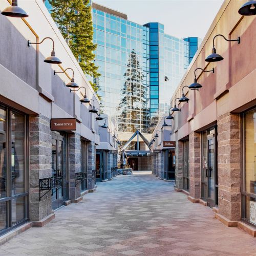 A sunny outdoor shopping arcade lined with storefronts like &ldquo;Double Diamond Jewelry,&rdquo; hanging lamps, and a glass office tower above.